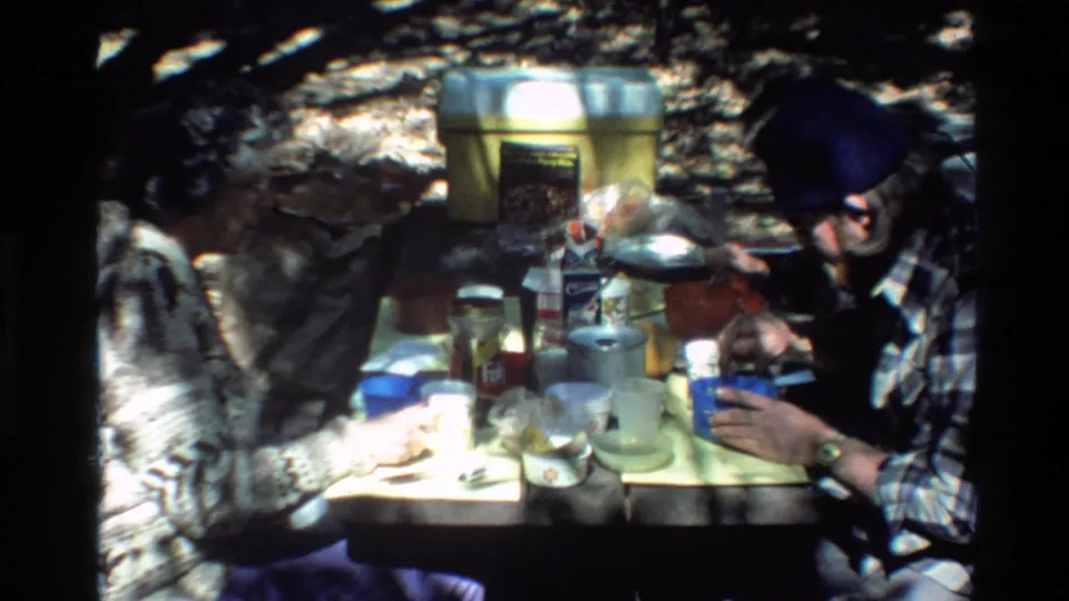 Big Sur 1980 home-movie frame showing two adults at a campsite table with dishes, cups, containers, and a coffee can.