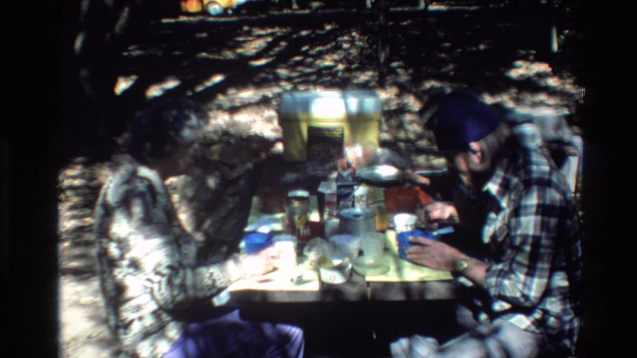 In 1980 Big Sur home-movie footage, two adult men in plaid shirts sit at a wooded picnic table with food, cups, and cookware.
