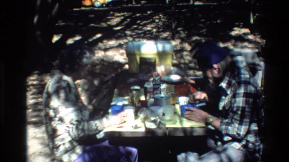 In 1980 Big Sur home-movie footage, two adult men in plaid shirts sit at a wooded picnic table with food, cups, and cookware.