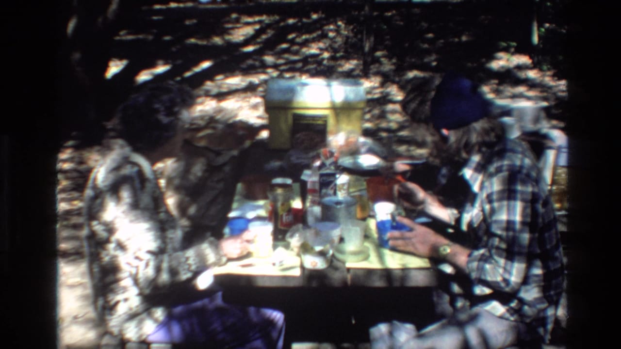 A bearded man in a dark beanie sits beside a picnic table covered with food items.