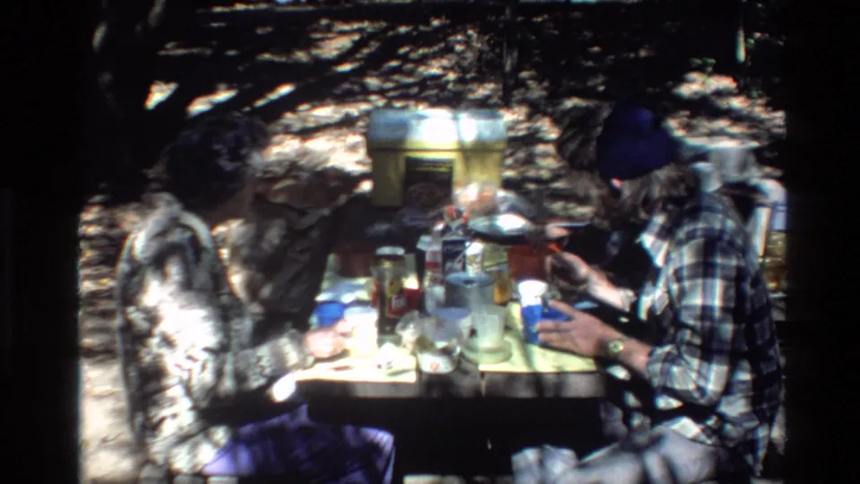 A bearded man in a dark beanie and plaid shirt sits at a Big Sur picnic table with cups, containers, and cookware in dappled light.