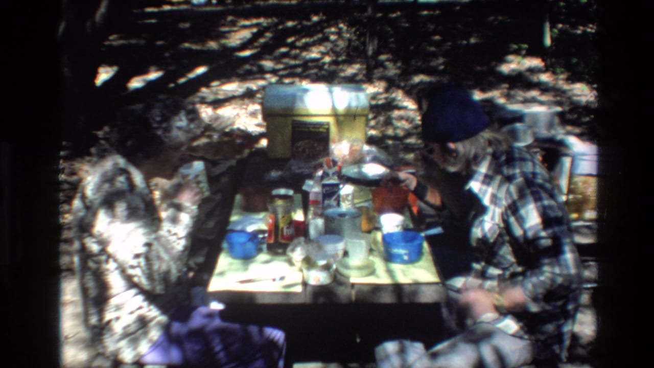 The home-movie frame shows two adult men at a wooded Big Sur picnic table, with plastic containers, cups, and scattered meal items visible.