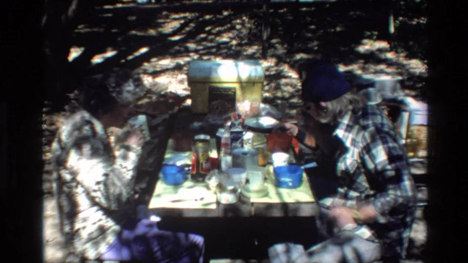 The home-movie frame shows two adult men at a wooded Big Sur picnic table, with plastic containers, cups, and scattered meal items visible.