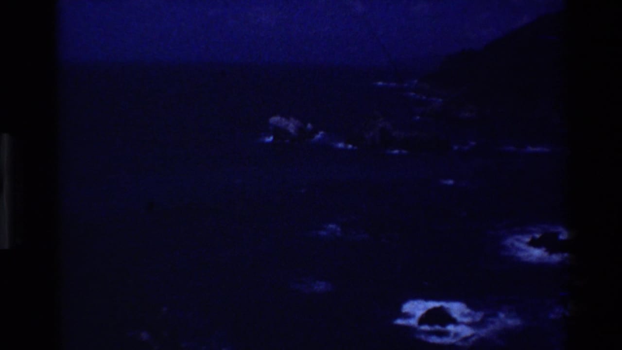 Underexposed 1980 Big Sur 8mm coastal view with blue water, rocky shoreline, and dark cliff shapes.