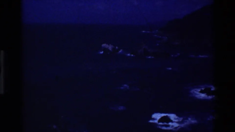 Underexposed 1980 Big Sur 8mm coastal view with blue water, rocky shoreline, and dark cliff shapes.