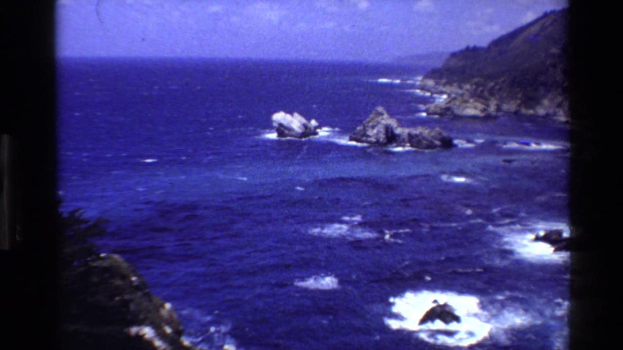 1980 Big Sur home-movie frame of deep blue Pacific water, rocky outcrops, breaking waves, and coastal cliffs.