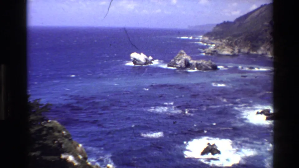 1980 8mm view of Big Sur, California, with deep blue water, white surf, offshore rocks, and coastal cliffs.