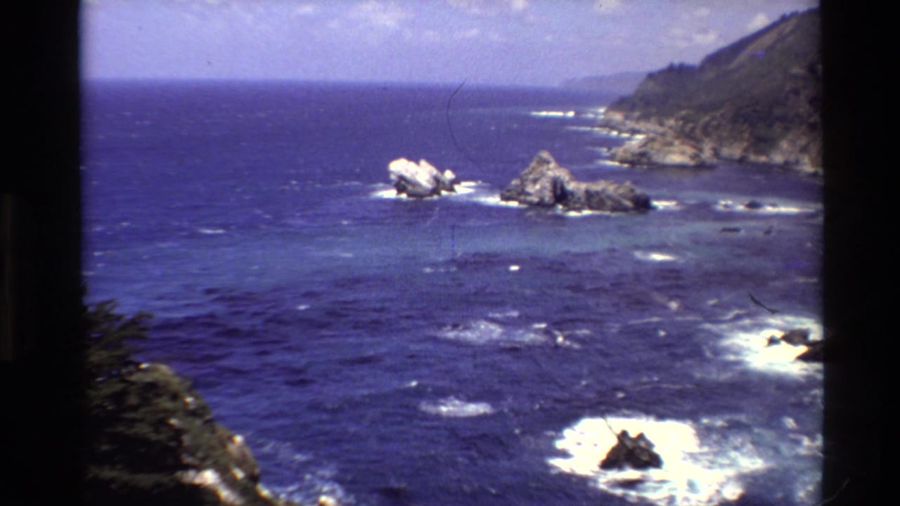 Rocky coastline and offshore formations sit in blue water with white wave breaks.