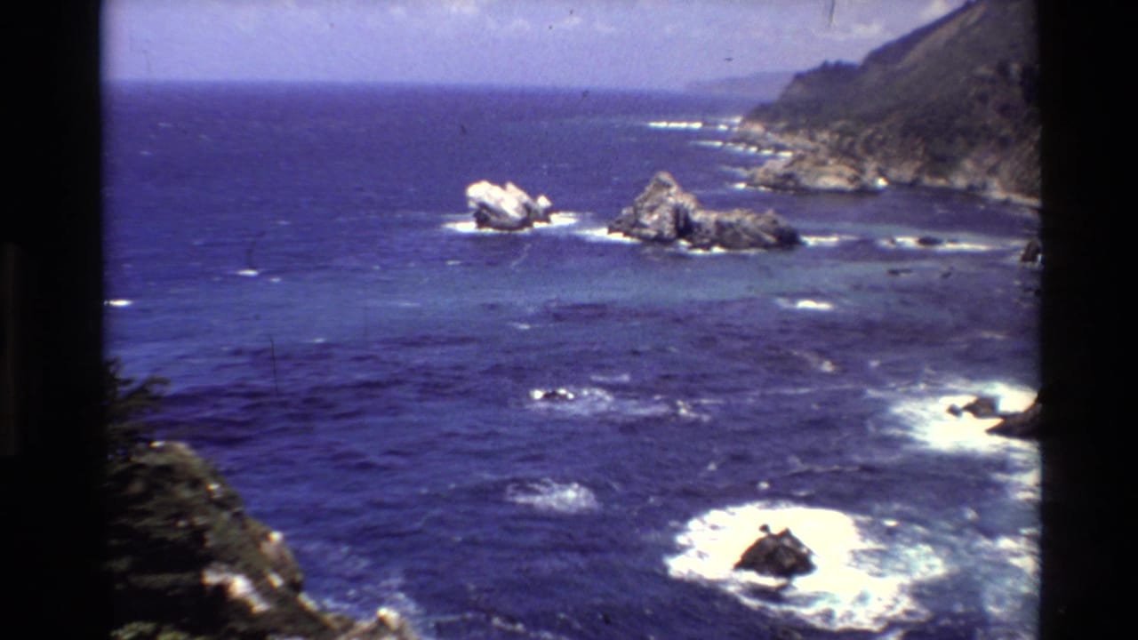 1980 8mm Big Sur coastal view with saturated blue ocean, foamy surf, offshore rocks, and rugged cliff shoreline.