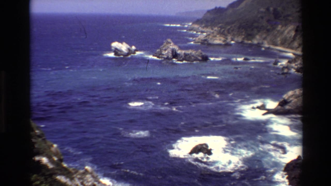Big Sur, California, in 1980 8mm footage, with white surf around nearshore rocks and a rugged coastal slope beyond.