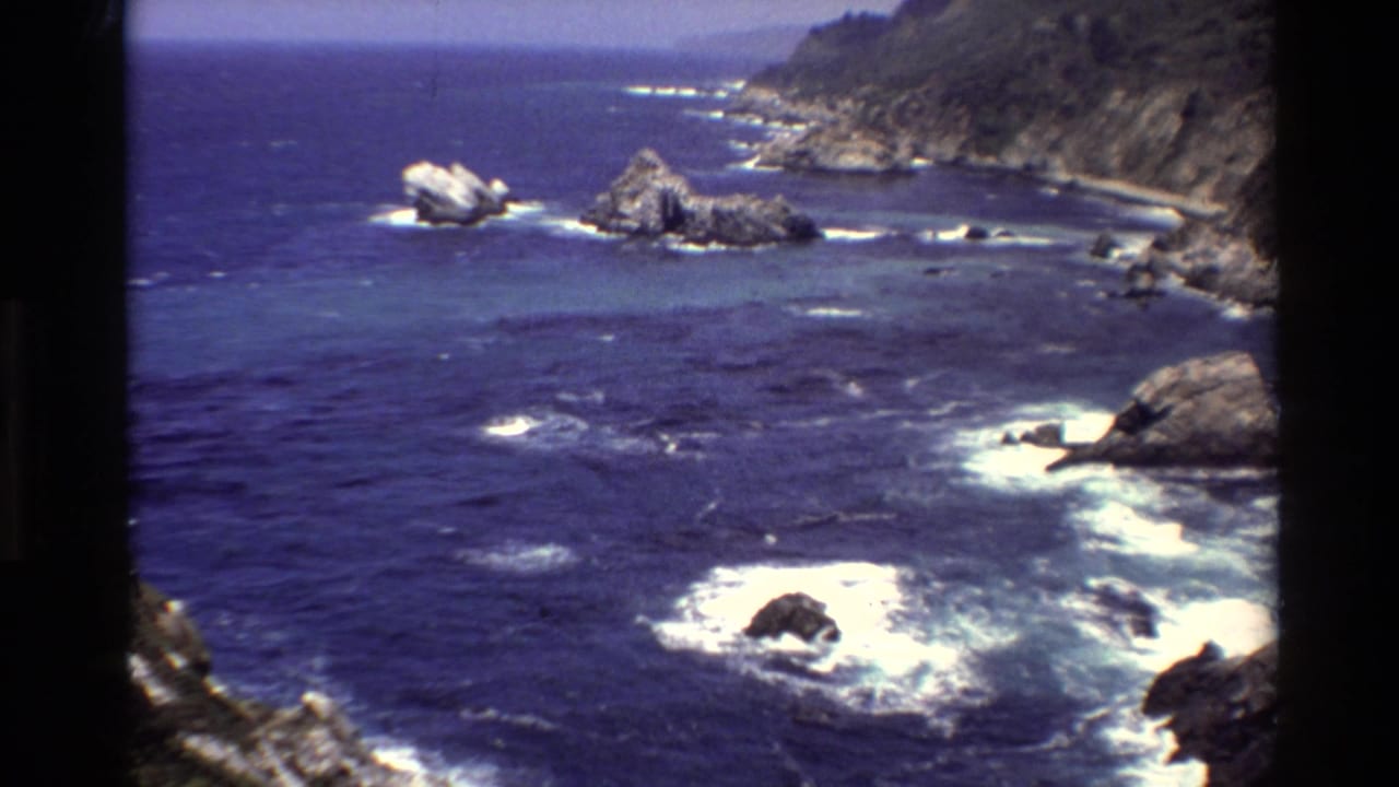 Blue ocean surf breaks around jagged rocks below a steep coastline.