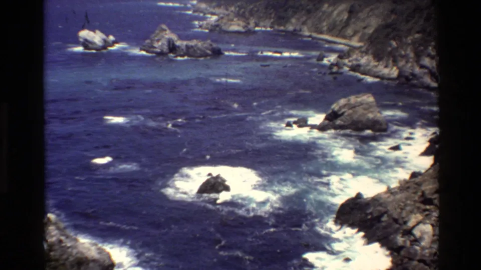 8mm Big Sur coastal view with saturated blue water, white foam, offshore rocks, and a rocky cliff edge.