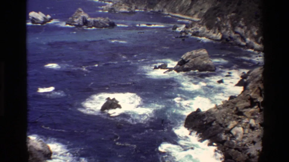 Vintage 8mm frame of Big Sur surf, with whitewater around jagged rocks and a steep Pacific shoreline in the background.