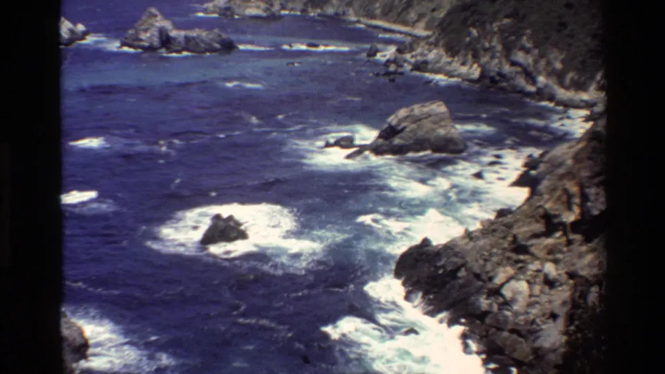1980 Big Sur 8mm frame showing blue Pacific water, foaming surf, offshore rocks, and a steep rocky shoreline.