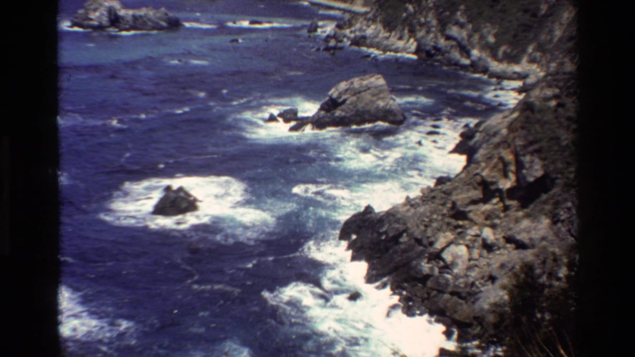 Ocean waves churn around dark rocks beside a steep rocky coast.