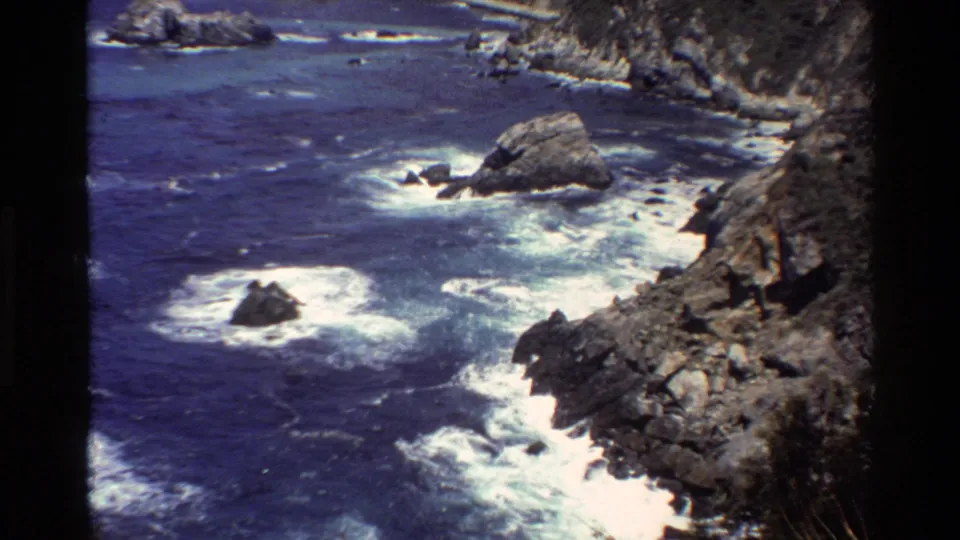 Big Sur coastal view with dark rocks, foaming blue water, and cliff edges in a grainy 8mm home-movie frame.