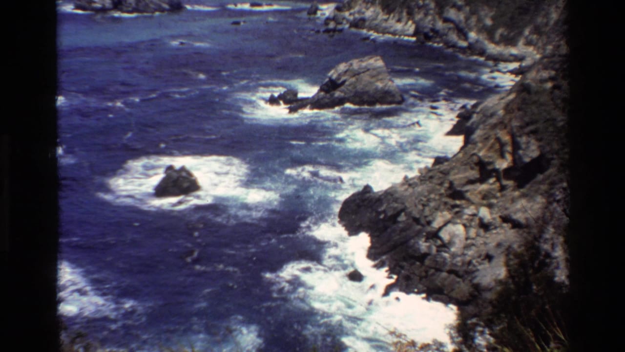 1980 Big Sur home-movie frame of a rugged shoreline, offshore rocks, and white surf spreading across deep blue water.