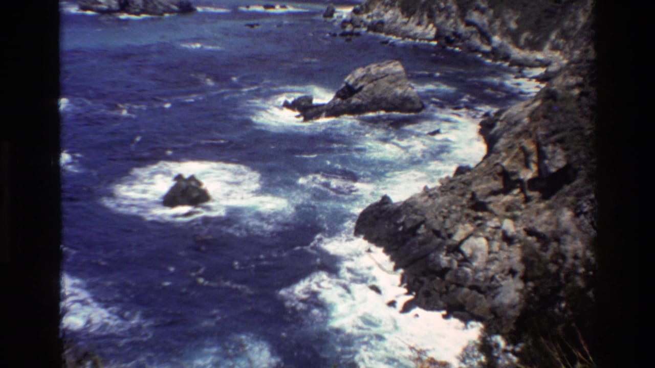 Grainy 8mm Big Sur coastal frame with blue water, white breakers, offshore rocks, and a steep rocky edge.