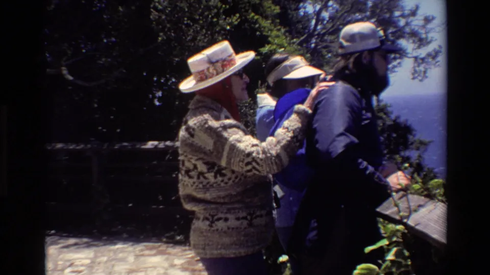 Two adults at a Big Sur coastal viewpoint in 1980 home-movie footage, one wearing a patterned sweater and broad hat beside another in a cap.