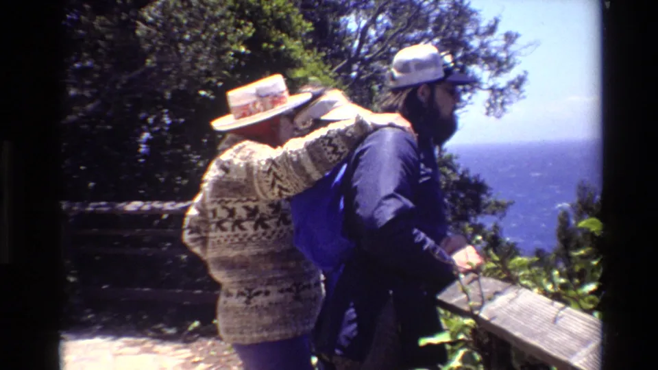 Home-movie frame of two adults at a Big Sur viewpoint, with a patterned sweater, blue backpack, railing, vegetation, and ocean in view.