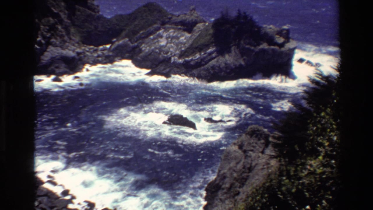 Elevated Big Sur ocean view showing foamy waves, offshore rocks, dark cliffs, and vegetation along the edge.