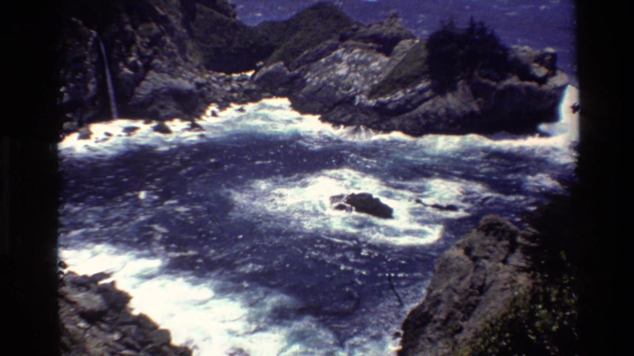 Choppy ocean water swirling around dark rocks below steep coastal cliffs.