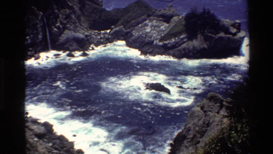 Big Sur shoreline seen on 8mm film, with white surf spreading through a rocky cove beneath rugged coastal slopes.