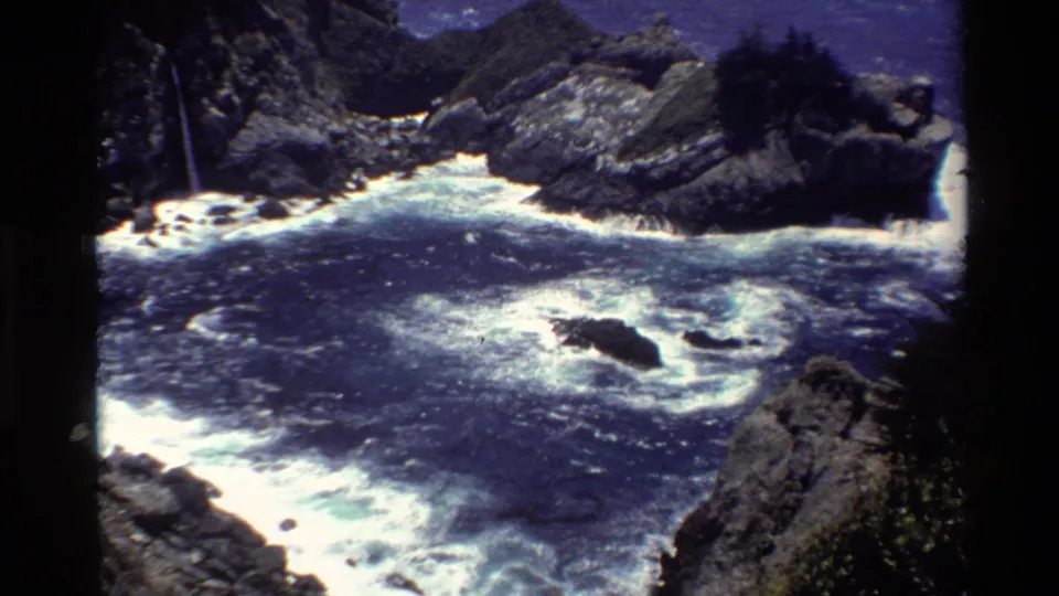 Vintage 8mm view of Big Sur surf wrapping around an offshore rock, with steep cliffs and a narrow waterfall visible on the left.