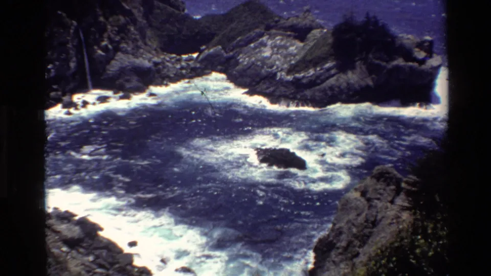 1980 Big Sur home-movie frame showing blue ocean, bright foam, and a dark rock below rugged cliffs and shadowed vegetation.