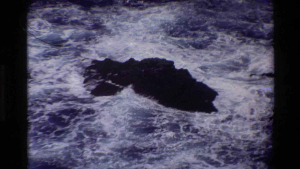 Foaming ocean waves surround a dark jagged rock offshore.