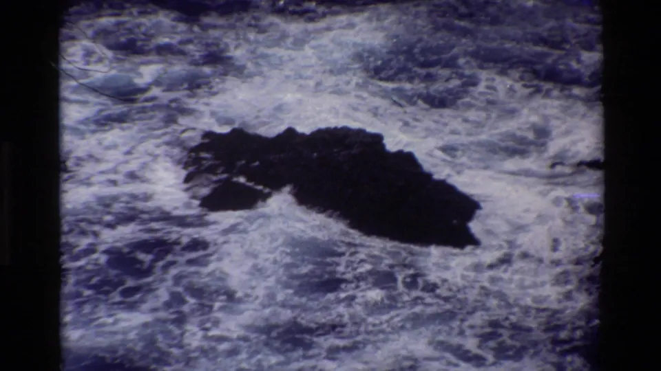 1980 Big Sur 8mm frame showing turbulent surf breaking around a dark rock in blue ocean water.