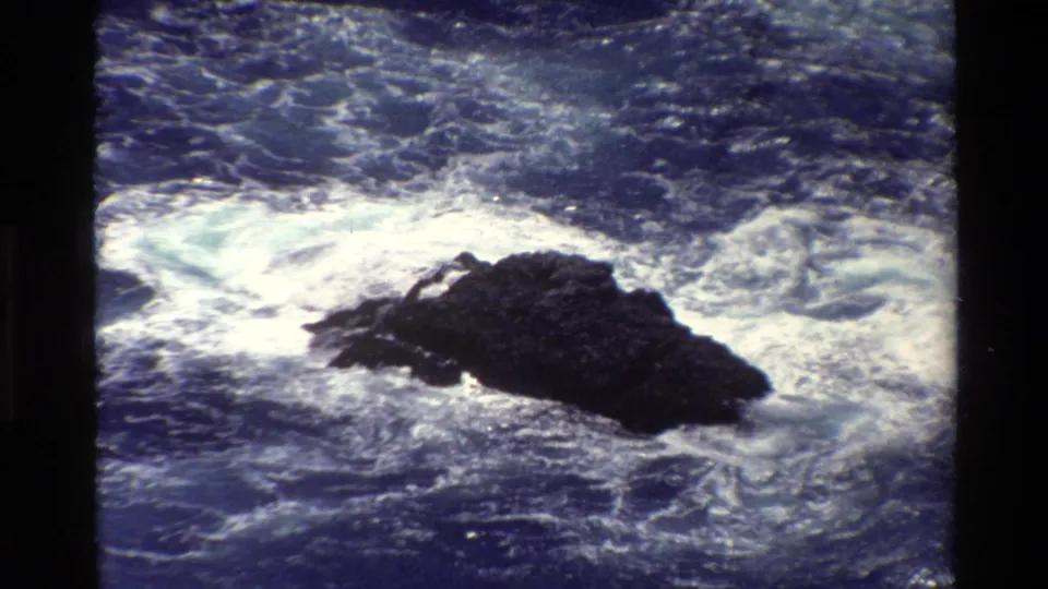 1980 Big Sur coastal frame with foaming waves wrapping around a dark, jagged offshore rock.