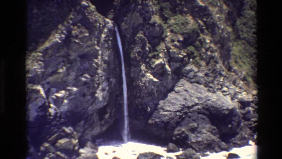 1980 Big Sur 8mm frame showing a thin waterfall falling through a rocky, moss-covered cliff area.