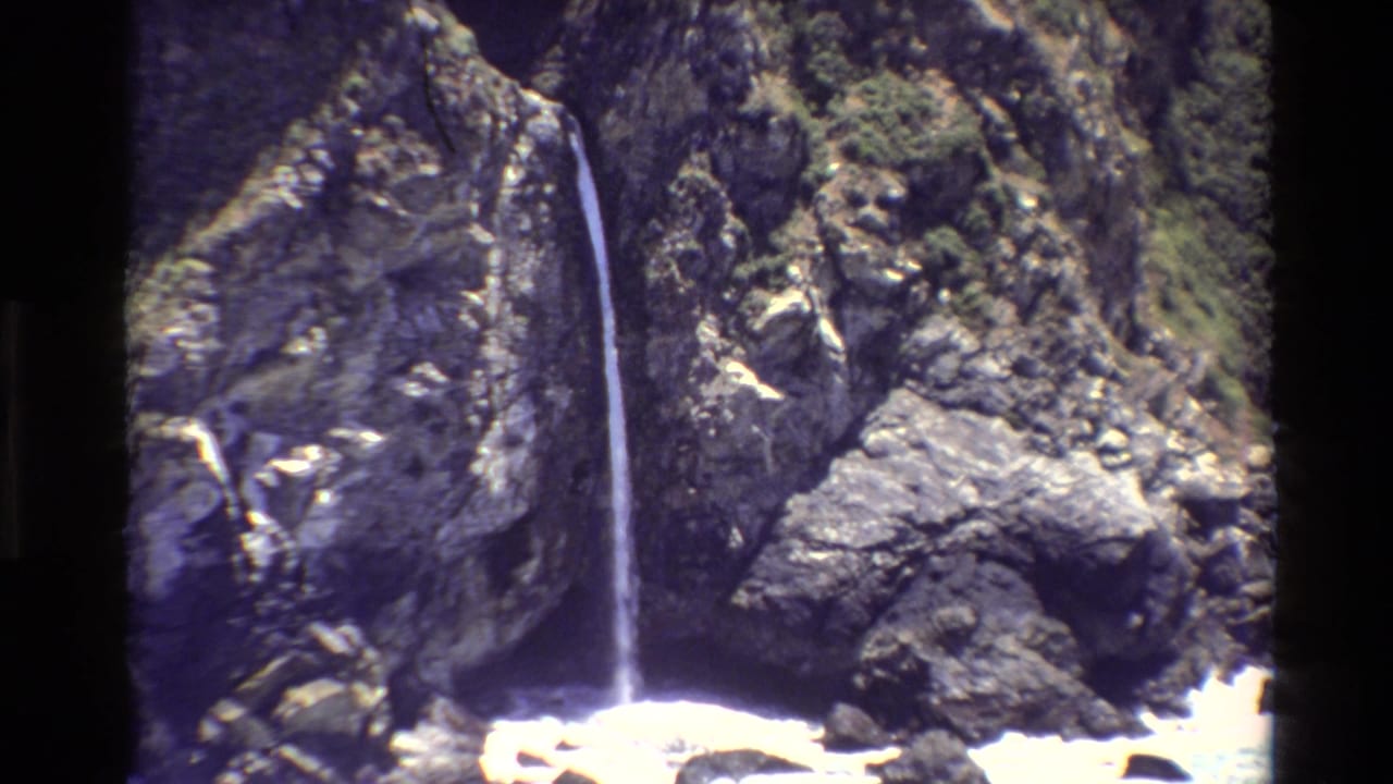 1980 Big Sur home-movie frame with a narrow waterfall set against steep rocks and patches of greenery.