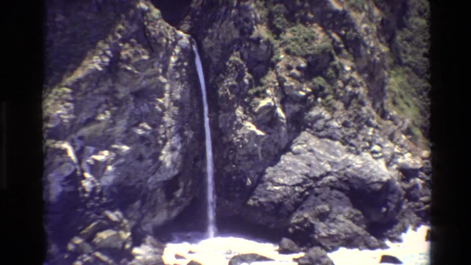 1980 Big Sur home-movie frame with a narrow waterfall set against steep rocks and patches of greenery.