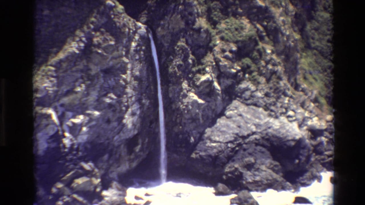 1980 Big Sur 8mm frame showing a thin waterfall descending a rugged cliff above white ocean foam.