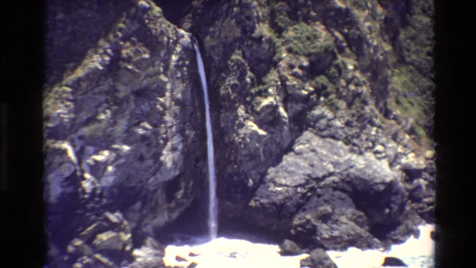 1980 Big Sur 8mm frame showing a thin waterfall descending a rugged cliff above white ocean foam.