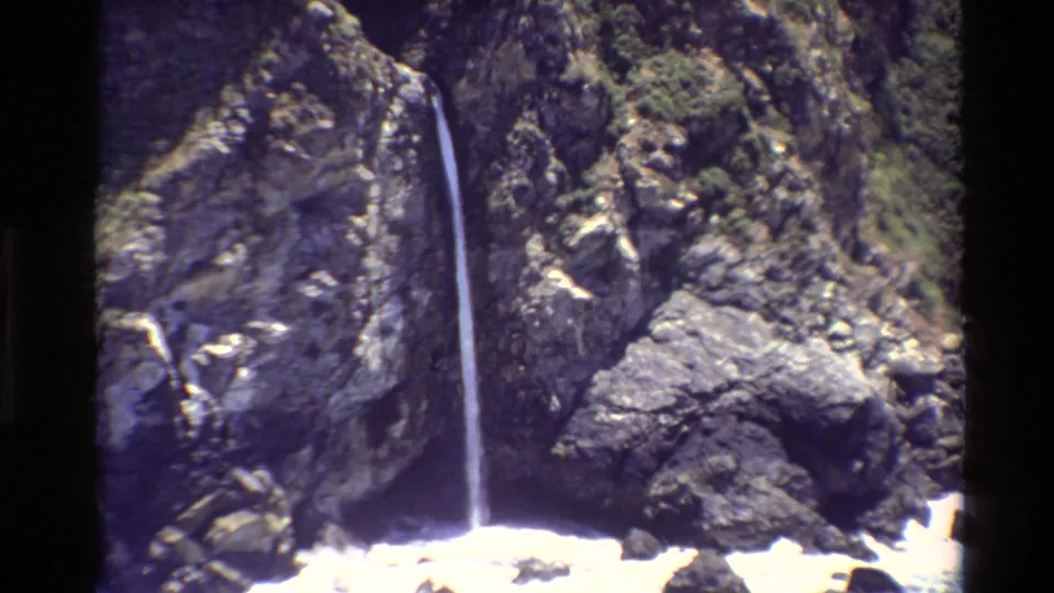 Big Sur coastal frame with a slender waterfall against dark rock and foaming water at the base.
