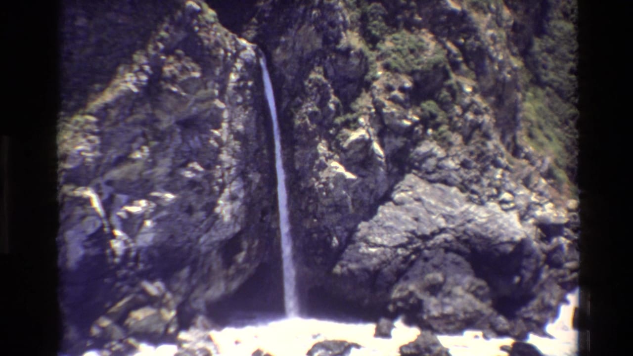 Vintage 8mm view of a Big Sur waterfall descending a textured cliff toward the ocean edge.