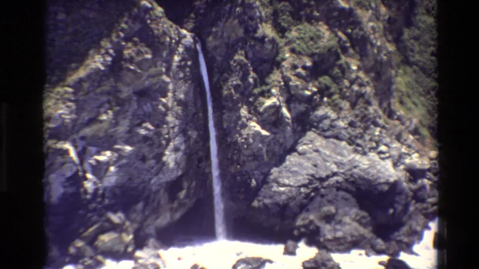Vintage 8mm view of a Big Sur waterfall descending a textured cliff toward the ocean edge.