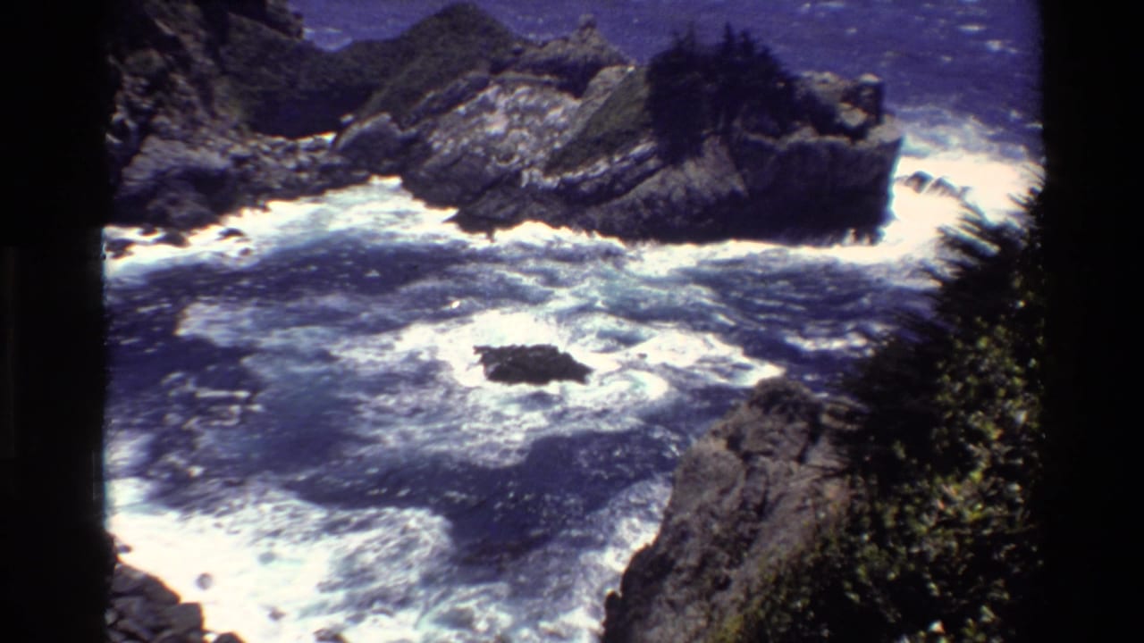 1980 Big Sur coastal frame showing turbulent blue water, white surf, jagged rocks, and dark cliff vegetation.