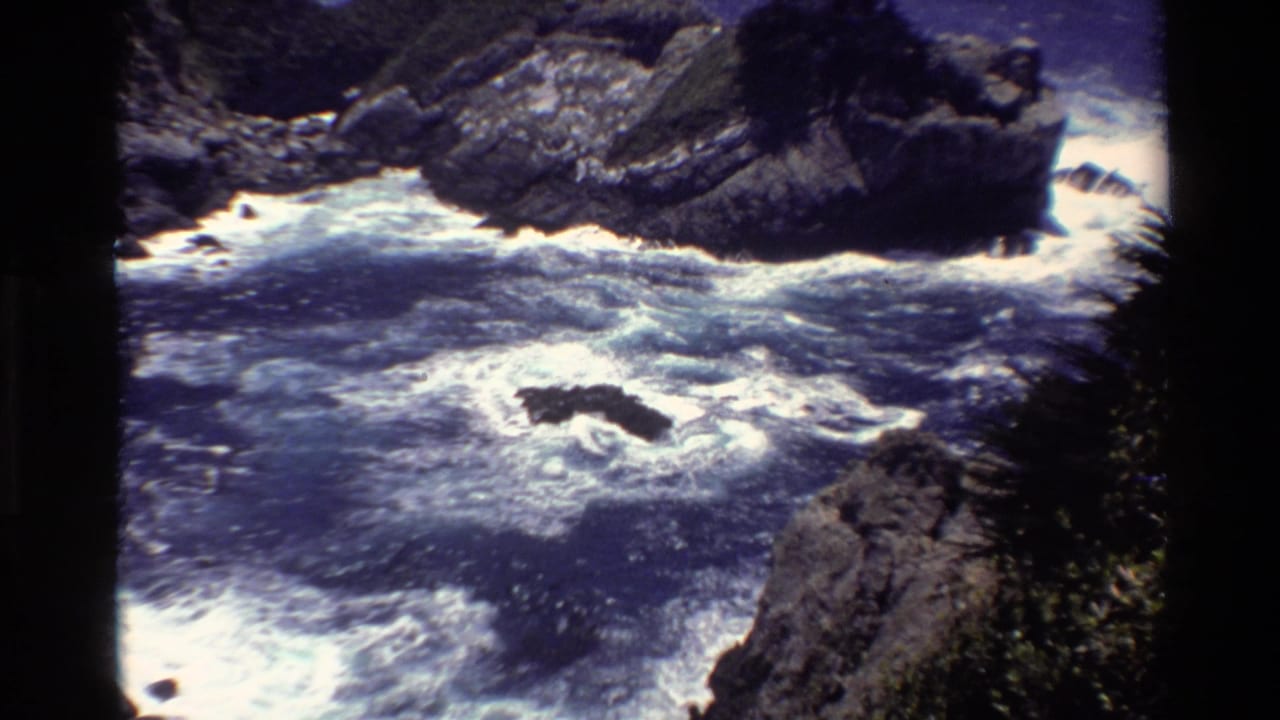 1980 Big Sur 8mm frame showing blue surf and white foam breaking around dark rocks near a steep shoreline.