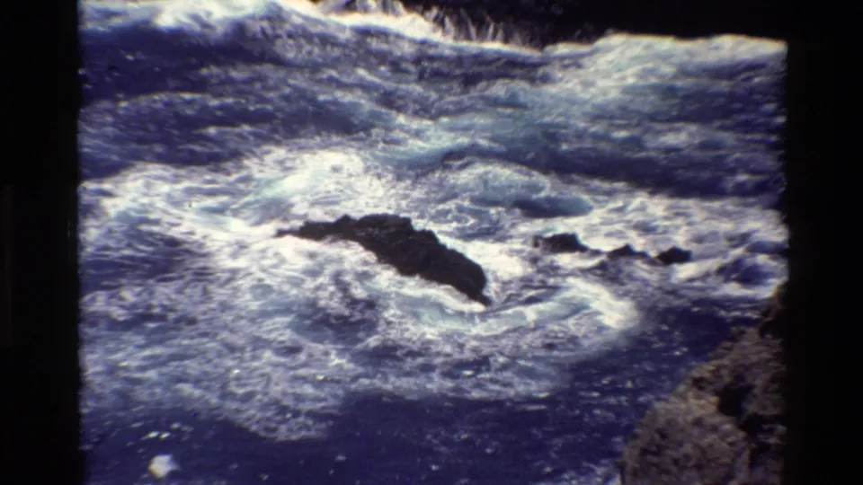 1980 Big Sur 8mm frame with turbulent surf wrapping around a dark rock outcrop in blue coastal water.