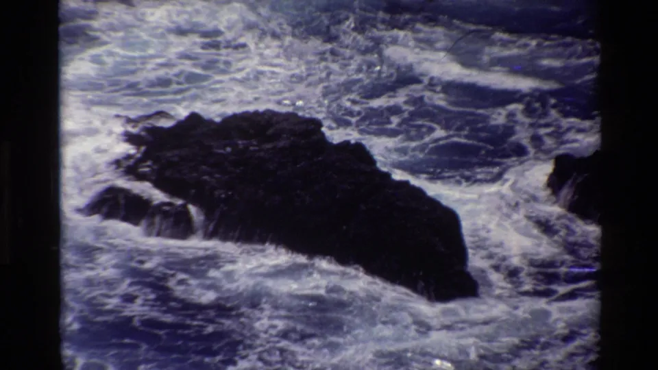 1980 Big Sur 8mm frame showing a large dark rock surrounded by white foam and rough coastal water.