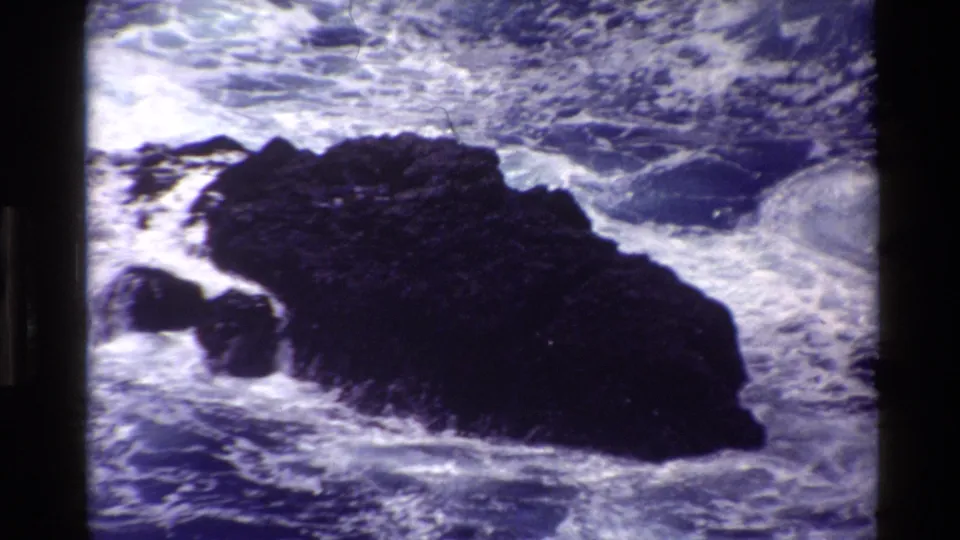 1980 Big Sur 8mm frame with a close view of foaming waves breaking around a dark coastal rock.