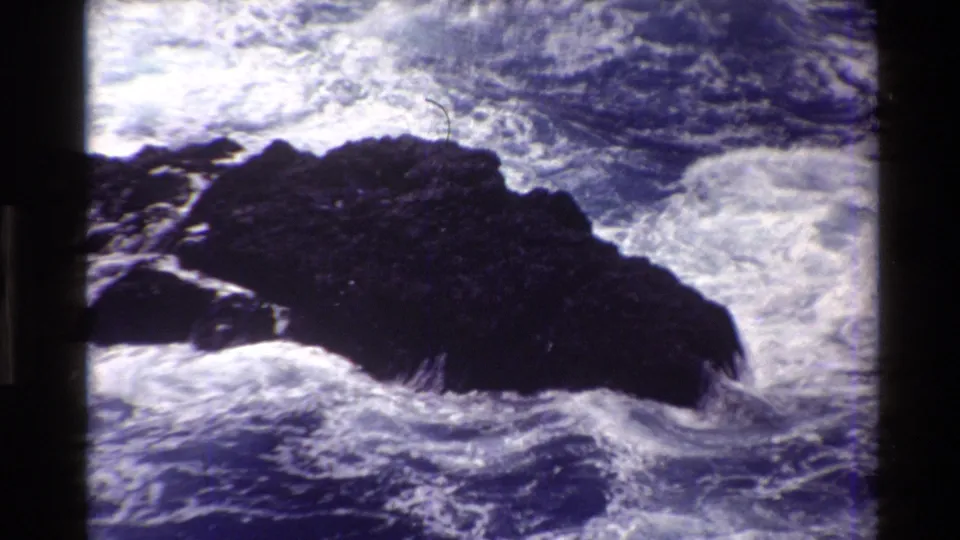 1980 Big Sur 8mm frame with turbulent blue water and white foam pushing against a dark coastal rock.