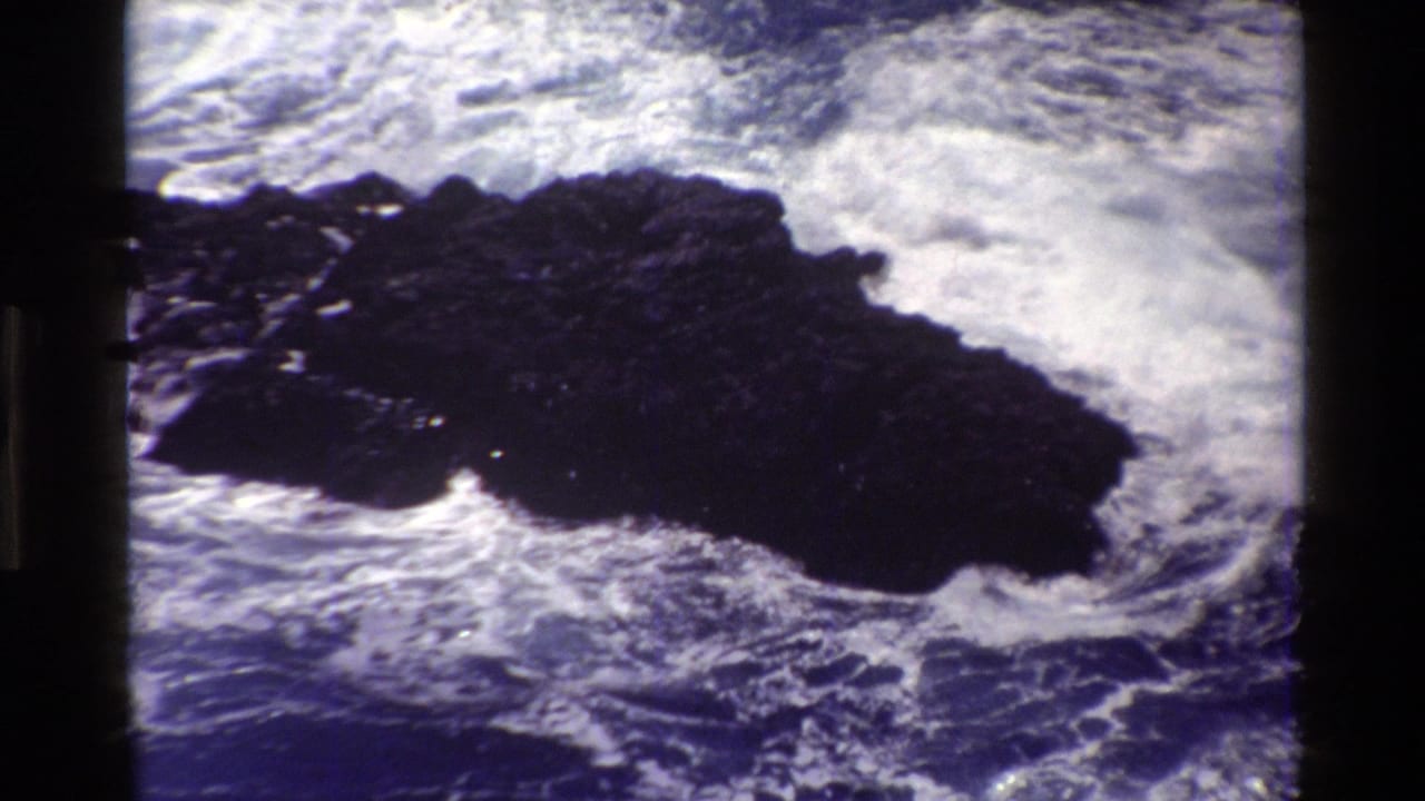 1980 Big Sur 8mm frame showing a dark shoreline rock with white Pacific surf breaking around it.