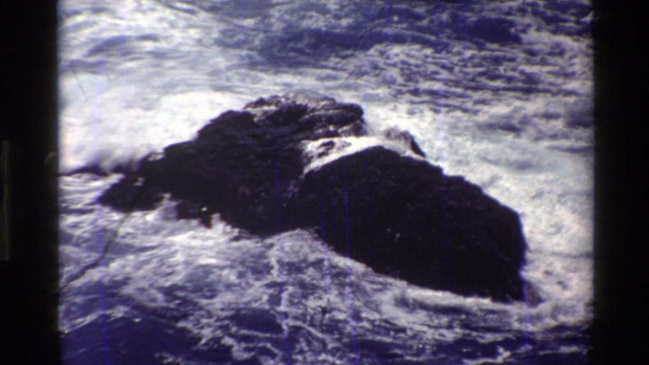 1980 Big Sur 8mm frame showing dark jagged rocks amid blue water and white breaking surf.
