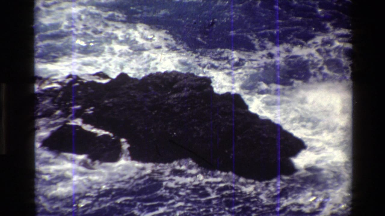1980 Big Sur 8mm frame showing white Pacific surf surrounding a dark jagged coastal rock under bright daylight.