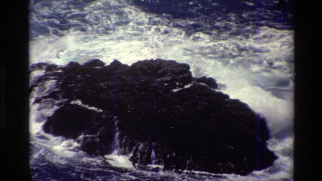 Big Sur coastal frame with dark rocks surrounded by turbulent white foam, recorded on grainy 8mm film.
